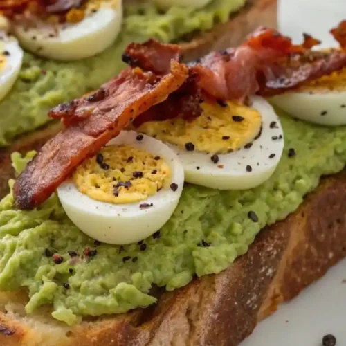 Avocado toast topped with creamy mashed avocado, sliced hard-boiled eggs, and crispy bacon strips on rustic bread.