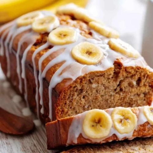 Moist banana bread topped with vanilla glaze and banana slices on a wooden table