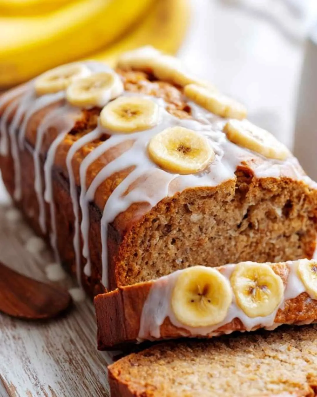Moist banana bread topped with vanilla glaze and banana slices on a wooden table