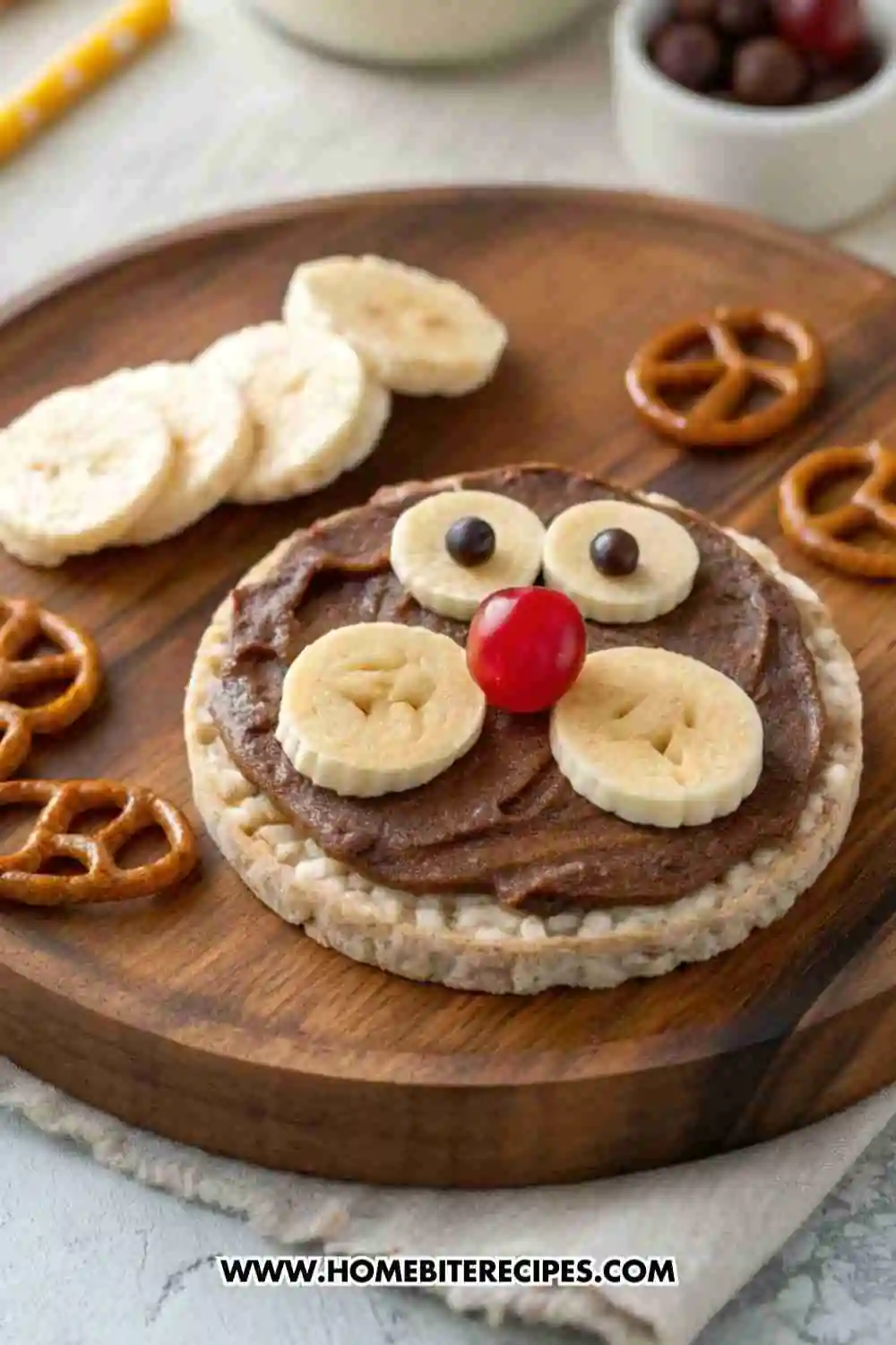 Bear face rice cake snack made with chocolate spread, banana slices, pretzels, and a cherry nose on a wooden board.