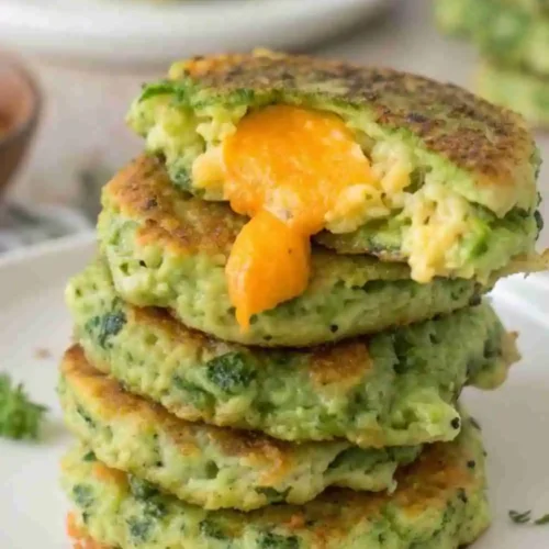 Stack of crispy cheesy broccoli fritters with melted cheddar cheese oozing out, served on a white plate.