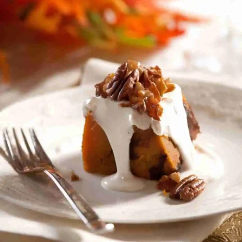 A slice of pumpkin pecan bread pudding topped with vanilla cream sauce and candied pecans, served on a white plate with autumn leaves in the background.