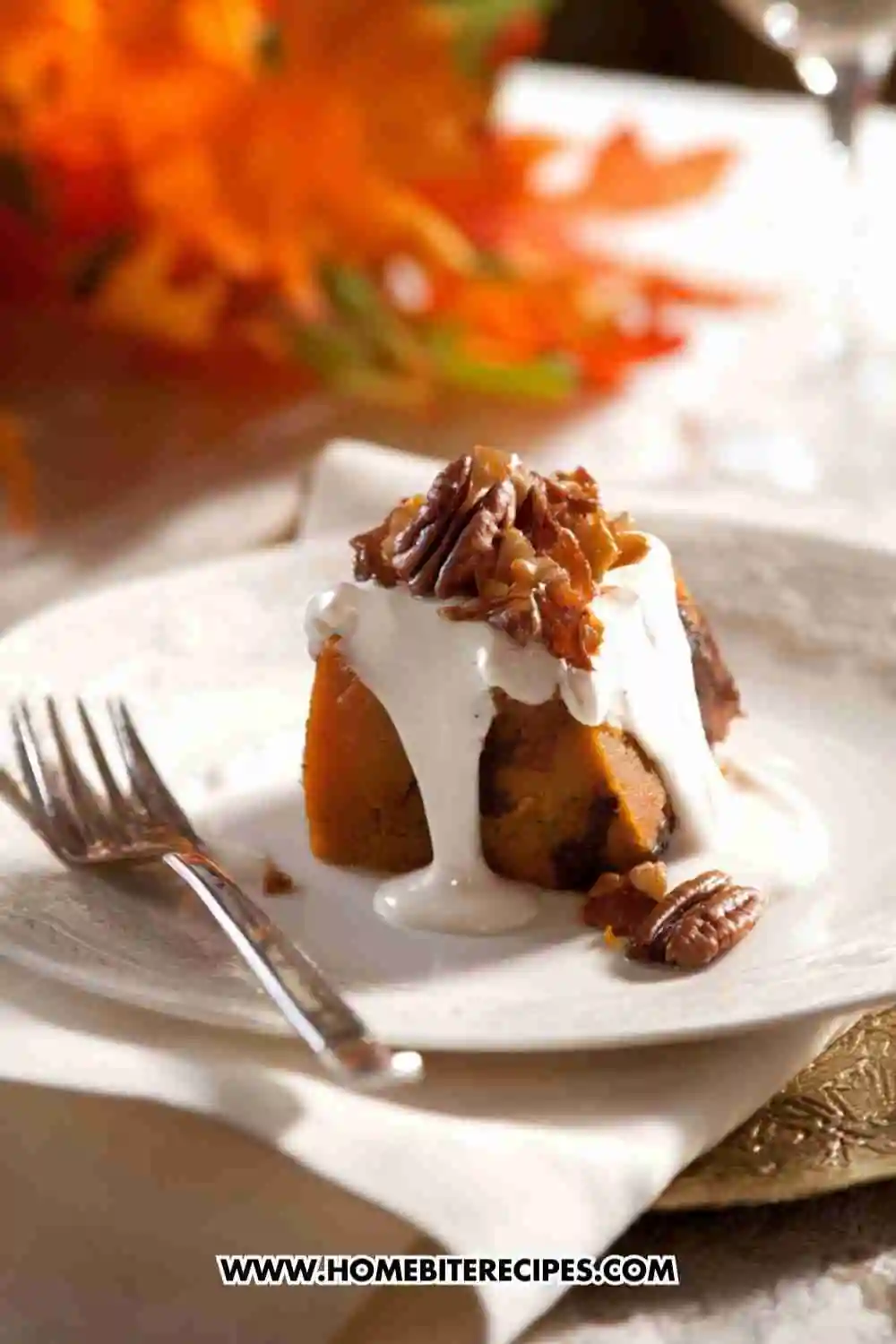 A slice of pumpkin pecan bread pudding topped with vanilla cream sauce and candied pecans, served on a white plate with autumn leaves in the background.