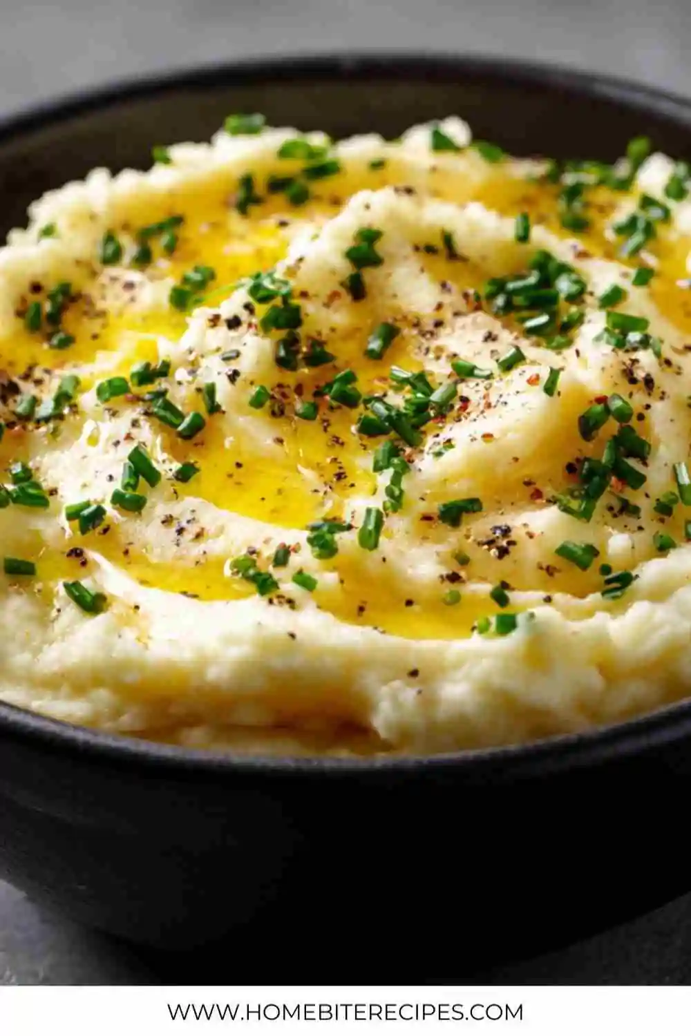 Smooth and creamy mashed potatoes topped with melted butter, freshly cracked black pepper, and chopped chives in a black bowl.