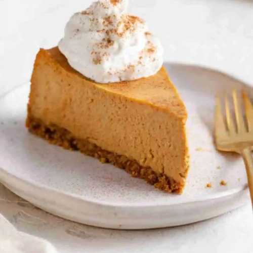 Slice of creamy baked pumpkin cheesecake with a gingersnap crust, topped with whipped cream and cinnamon on a white plate.