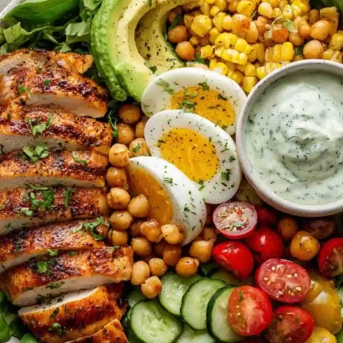 Grilled chicken power bowl with avocado, chickpeas, hard-boiled eggs, corn, cucumbers, tomatoes, and herb dressing.