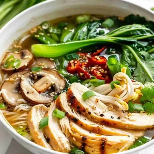 A steaming bowl of healthy chicken ramen noodle soup with sliced chicken breast, mushrooms, bok choy, chili slices, and green onions in a flavorful broth.