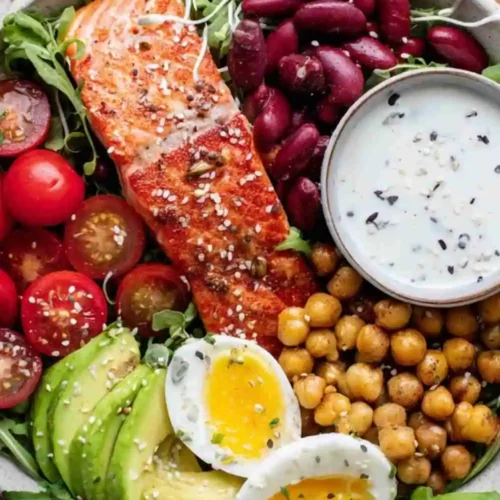 A vibrant salmon power bowl with grilled salmon, avocado, chickpeas, kidney beans, cherry tomatoes, soft-boiled egg, and yogurt dressing.