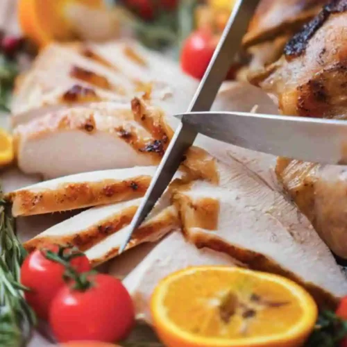 Juicy roast turkey being sliced with fresh herbs, oranges, and tomatoes on a festive platter.