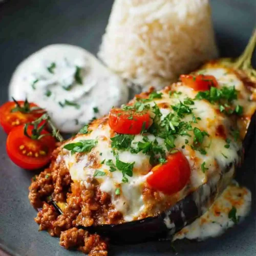 Baked Mediterranean stuffed eggplant filled with ground beef, rice, and tomato sauce, topped with melted cheese, cherry tomatoes, and parsley on a plate with yogurt sauce and rice.