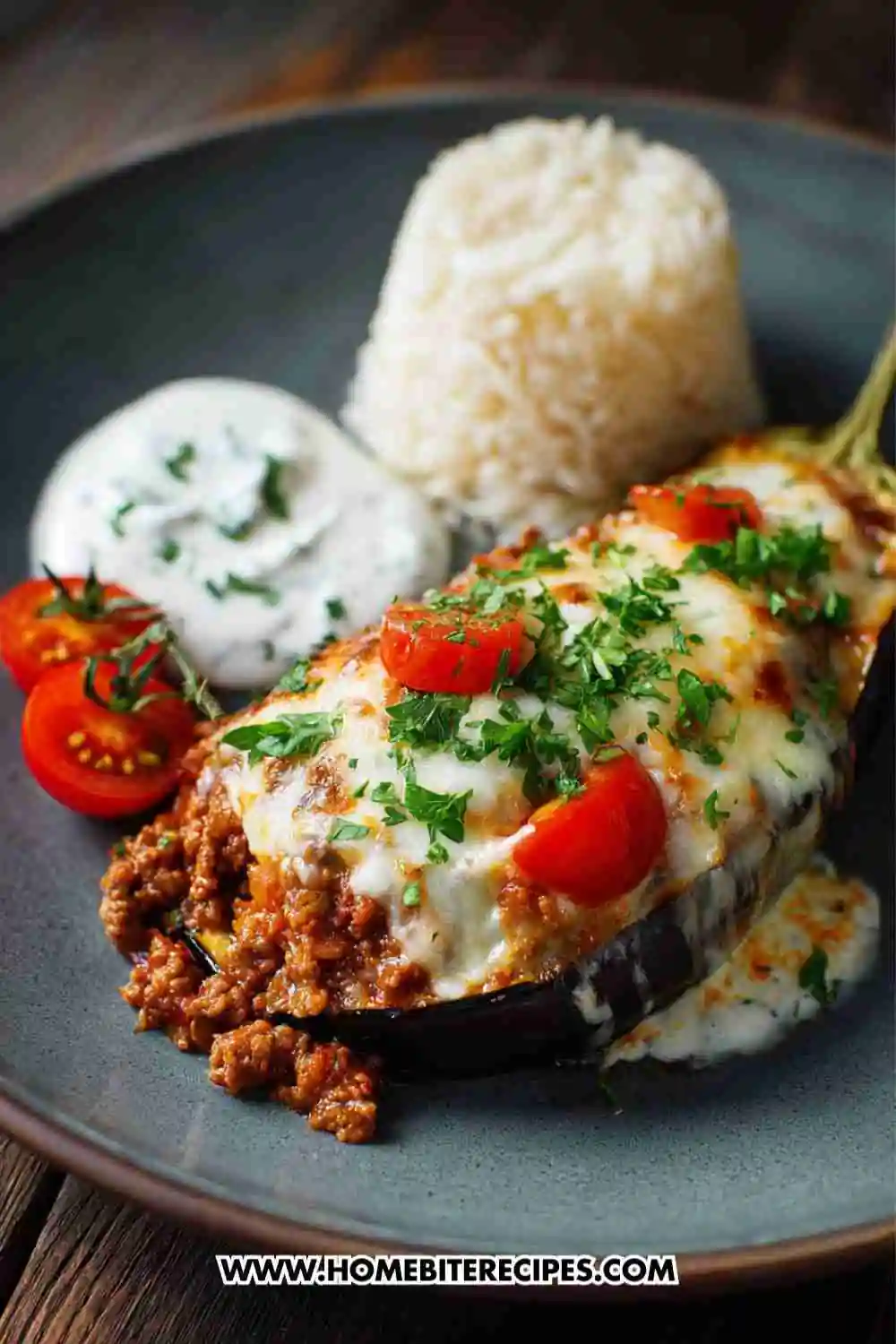 Baked Mediterranean stuffed eggplant filled with ground beef, rice, and tomato sauce, topped with melted cheese, cherry tomatoes, and parsley on a plate with yogurt sauce and rice.