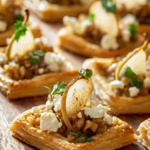 Mini puff pastry bites topped with caramelized pear slices, creamy goat cheese, walnuts, and herbs on a wooden board.