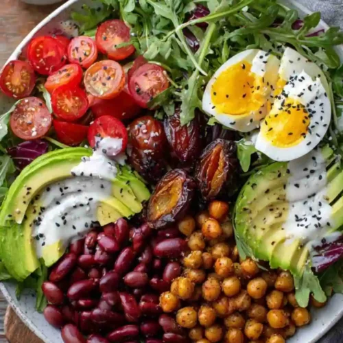 Colorful salad bowl with crispy chickpeas, avocado slices, boiled eggs, cherry tomatoes, dates, red beans, and yogurt dressing on arugula greens.