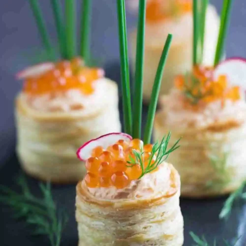 Elegant puff pastry appetizers filled with creamy salmon mousse, topped with orange caviar, fresh dill, and chives, served on a dark slate board.