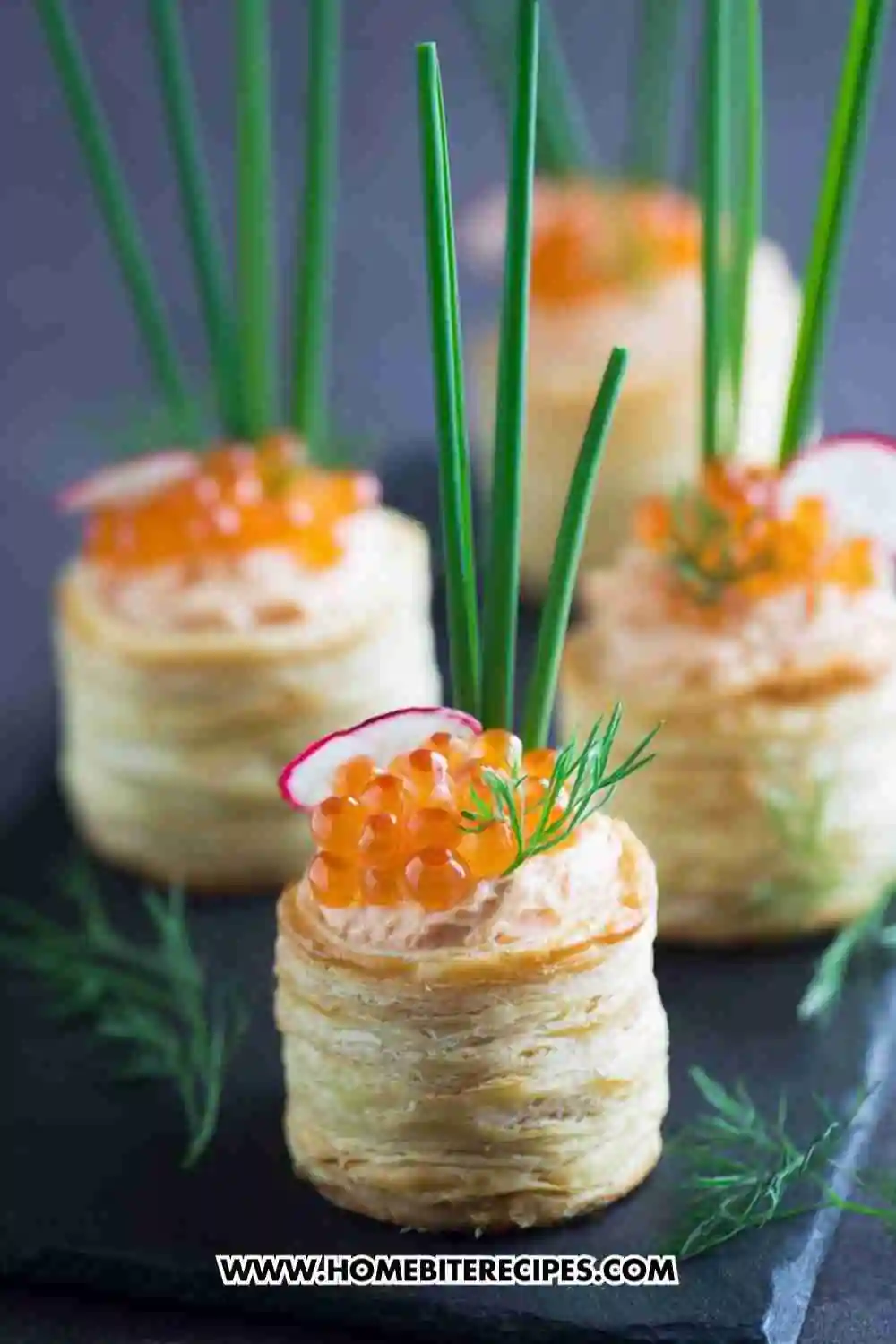Elegant puff pastry appetizers filled with creamy salmon mousse, topped with orange caviar, fresh dill, and chives, served on a dark slate board.