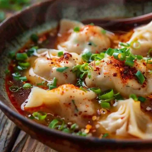A bowl of spicy dumpling soup with chili oil broth, green onions, and tender dumplings served hot and steaming.