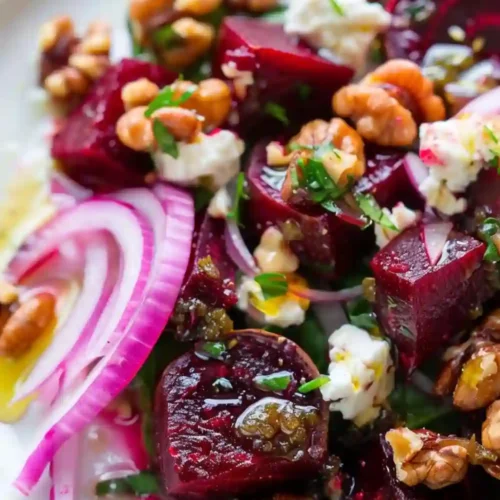 Close-up of a beetroot salad with roasted beet cubes, feta cheese, walnuts, red onion slices, herbs, and olive oil served on a white plate.