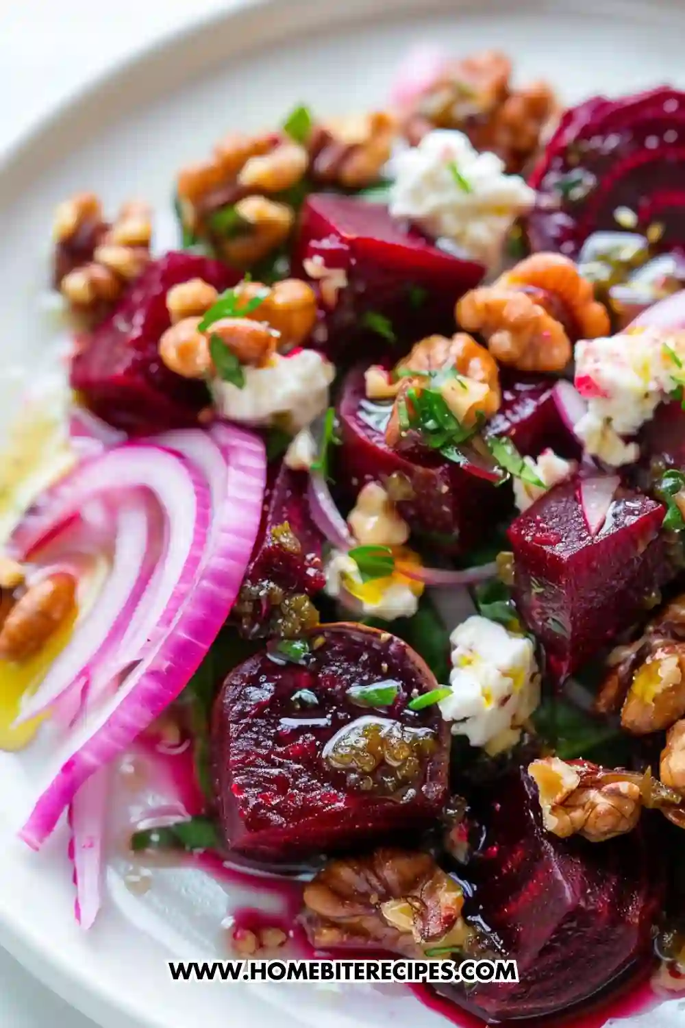 Close-up of a beetroot salad with roasted beet cubes, feta cheese, walnuts, red onion slices, herbs, and olive oil served on a white plate.
