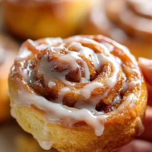 Close-up image of a warm, freshly baked apple cinnamon roll topped with creamy vanilla icing being held by a hand, with more cinnamon rolls blurred in the background.