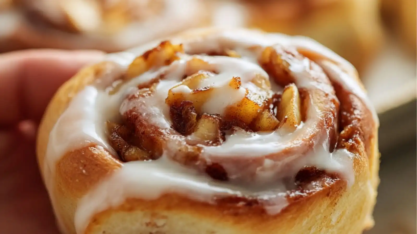 Close-up image of a warm, freshly baked apple cinnamon roll topped with creamy vanilla icing being held by a hand, with more cinnamon rolls blurred in the background.