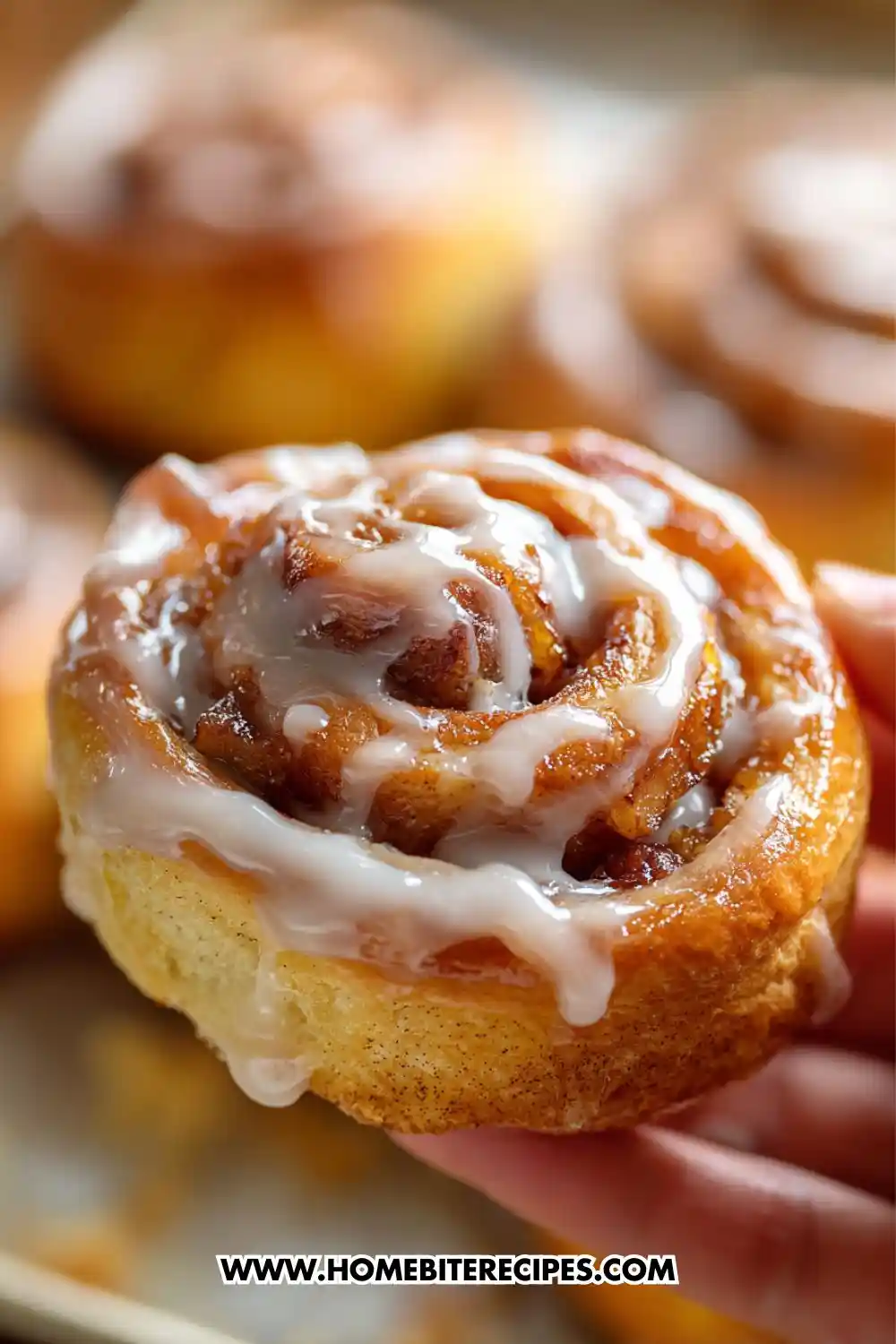 Close-up image of a warm, freshly baked apple cinnamon roll topped with creamy vanilla icing being held by a hand, with more cinnamon rolls blurred in the background.