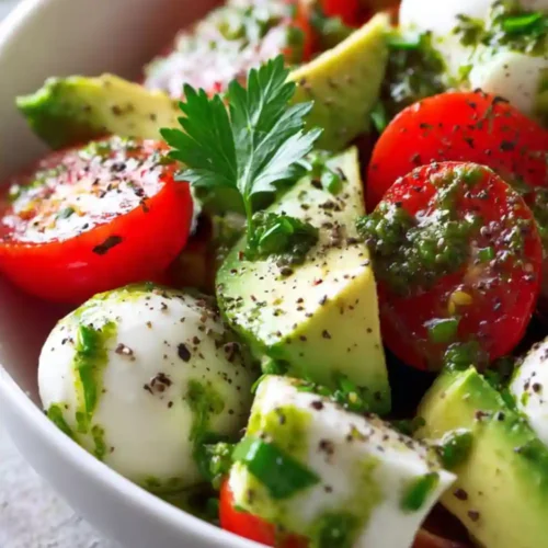 Colorful bowl of Avocado Caprese Salad with Fresh Pesto Dressing made with cherry tomatoes, mozzarella balls, avocado slices, and pesto dressing, garnished with fresh parsley.