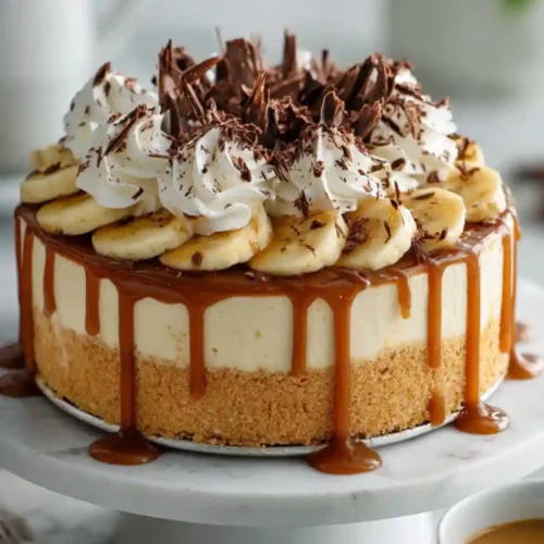 A rich no-bake banoffee cheesecake topped with caramel drizzle, fresh banana slices, whipped cream swirls, and chocolate shavings on a marble cake stand.
