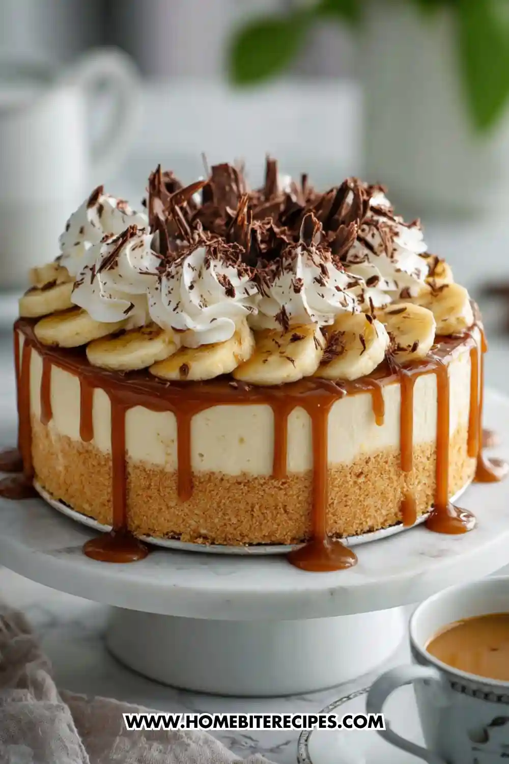 A rich no-bake banoffee cheesecake topped with caramel drizzle, fresh banana slices, whipped cream swirls, and chocolate shavings on a marble cake stand.