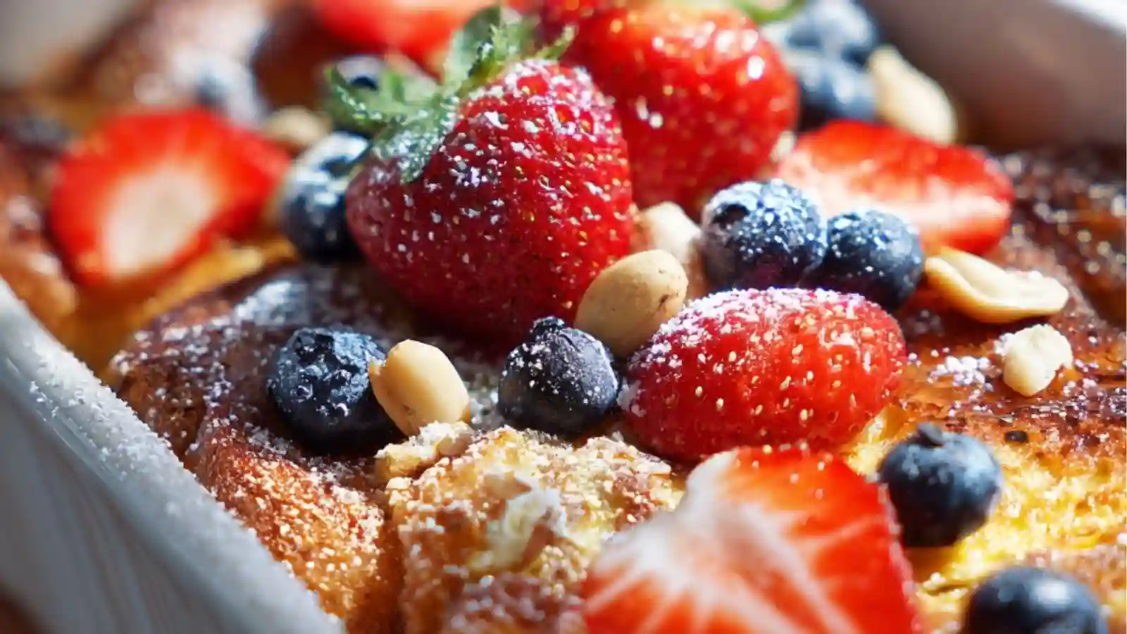A close-up of a baked French toast casserole topped with fresh blueberries, sliced strawberries, nuts, and powdered sugar in a white baking dish.