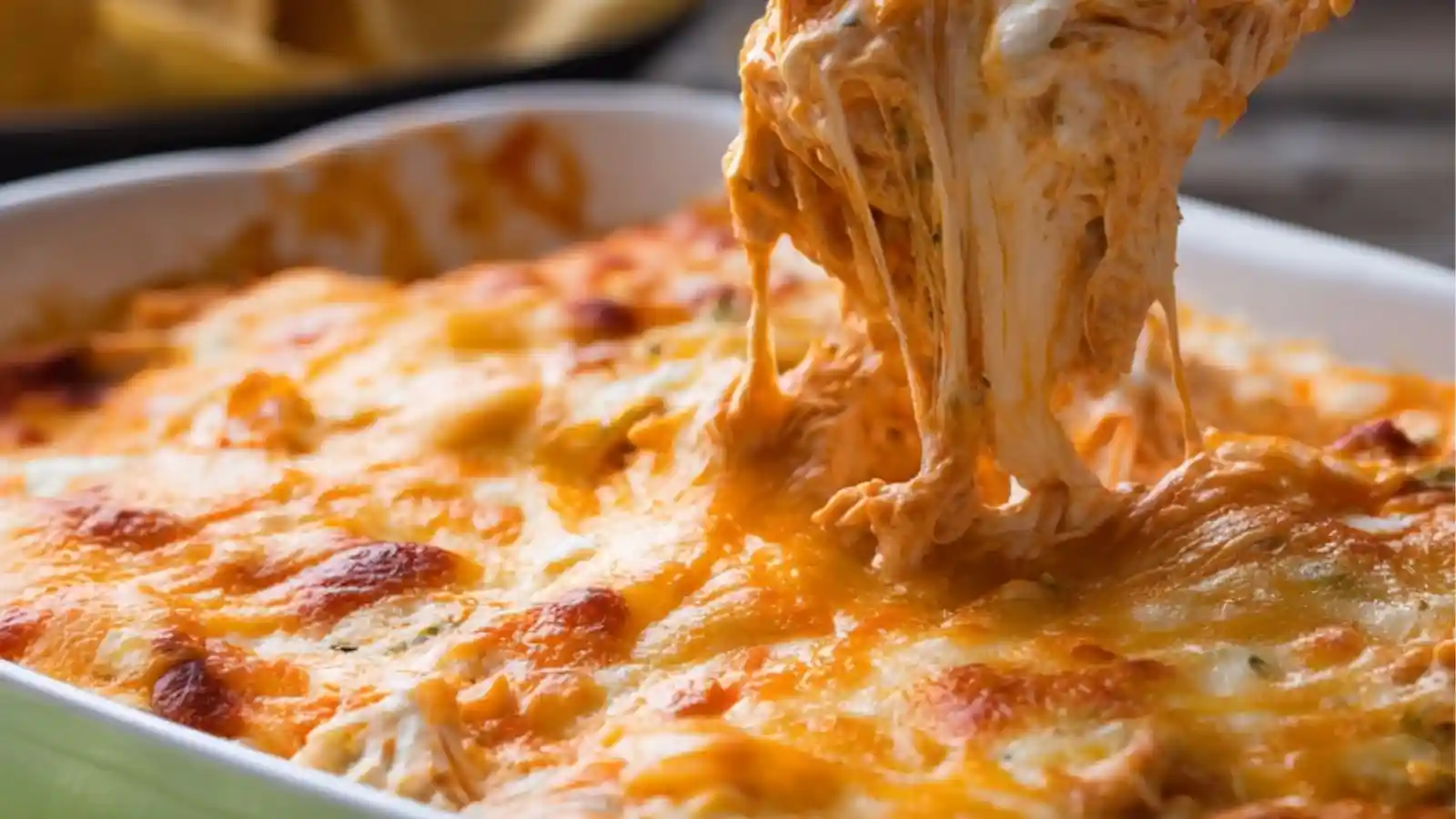 A hand dipping a tortilla chip into a hot, cheesy buffalo chicken dip topped with melted cheddar and green onions in a white baking dish.