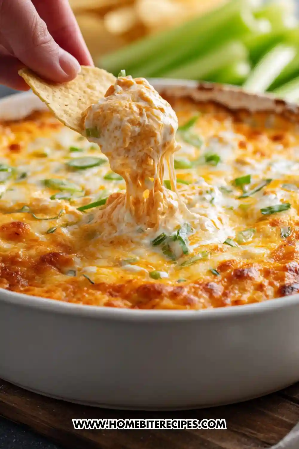 A hand dipping a tortilla chip into a hot, cheesy buffalo chicken dip topped with melted cheddar and green onions in a white baking dish.