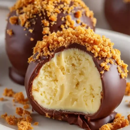 Close-up of chocolate-covered cheesecake bites coated with cookie crumbs, with one bite showing the creamy cheesecake center.
