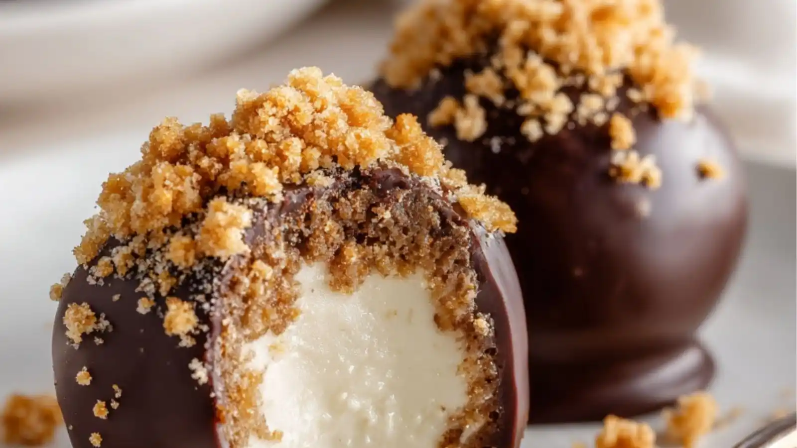 Close-up of chocolate-covered cheesecake bites coated with cookie crumbs, with one bite showing the creamy cheesecake center.