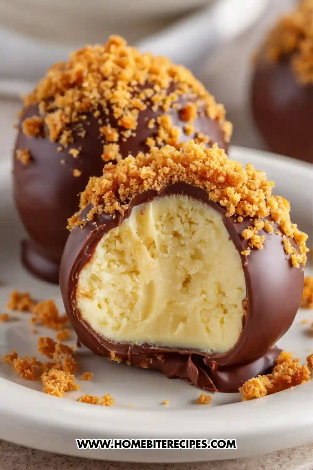 Close-up of chocolate-covered cheesecake bites coated with cookie crumbs, with one bite showing the creamy cheesecake center.