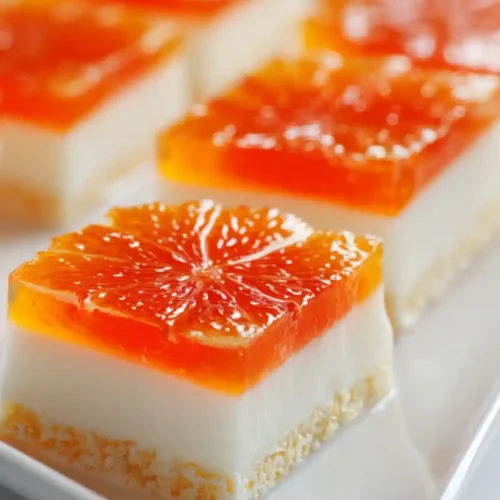A close-up of layered citrus dessert bites featuring a bright orange gelatin top with citrus slices, creamy white middle layer, and a crumbly biscuit base, served neatly on a white platter.