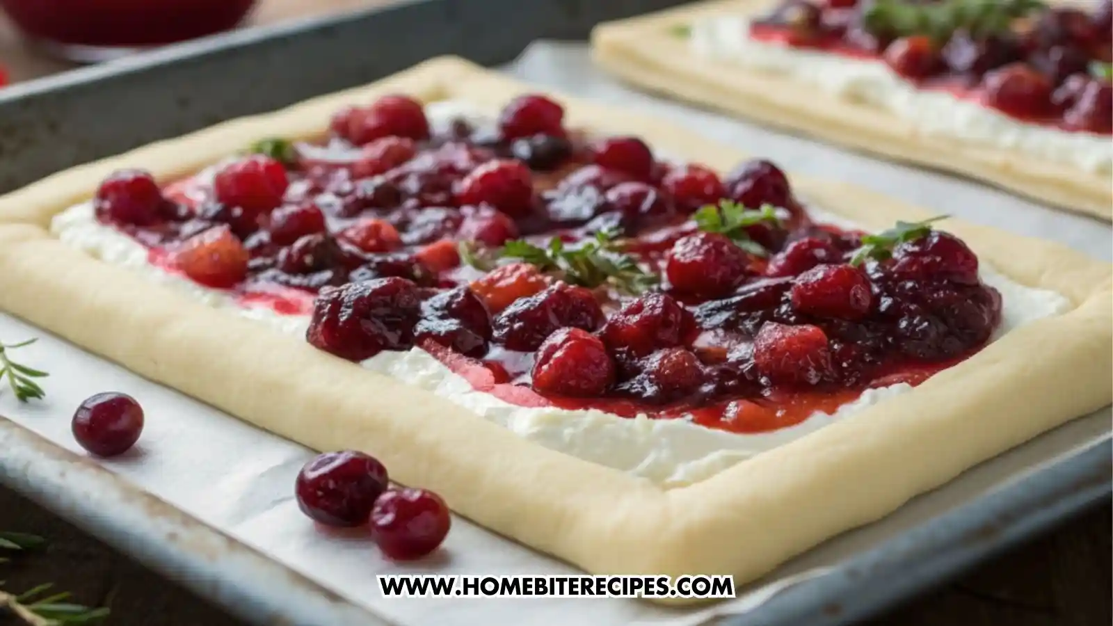 A close-up of raw puff pastry spread with goat cheese, topped with cranberries and cranberry sauce, all placed on a baking sheet.
