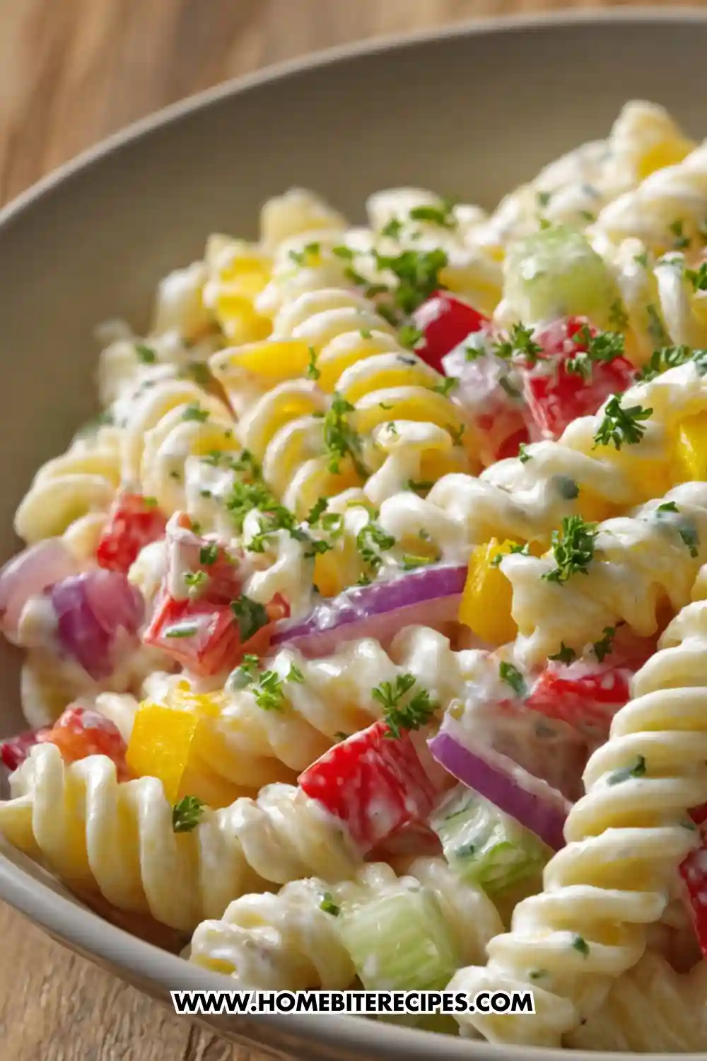 A bowl of creamy rotini pasta salad mixed with colorful vegetables such as red bell peppers, yellow peppers, cucumber, and red onion, topped with fresh parsley.