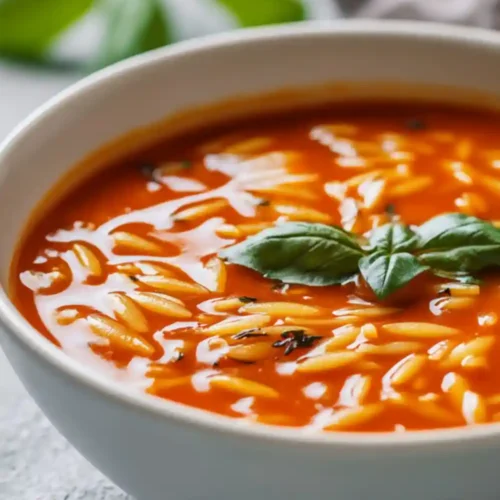 A bowl of creamy tomato orzo soup garnished with fresh basil leaves, served in a white bowl on a dark background.