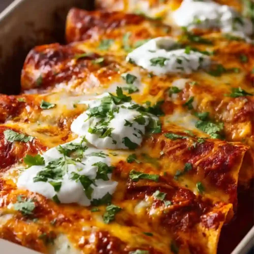 Close-up image of cheesy chicken enchiladas baked in a casserole dish, topped with melted cheese, red enchilada sauce, dollops of sour cream, and fresh chopped cilantro.