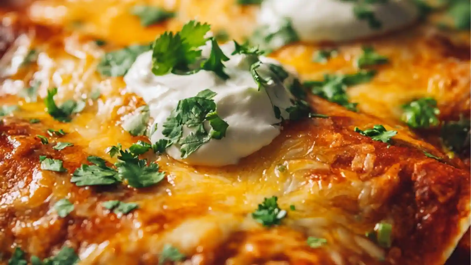 Close-up image of Easy Cheesy Chicken Enchiladas baked in a casserole dish, topped with melted cheese, red enchilada sauce, dollops of sour cream, and fresh chopped cilantro.