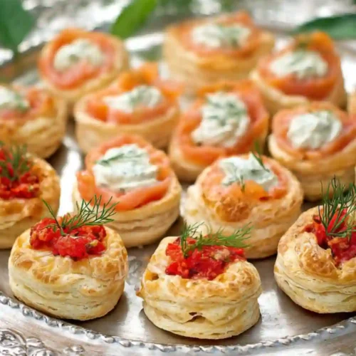 Mini puff pastry vol-au-vents filled with smoked salmon, herbed cream cheese, and red pepper spread, arranged on a silver platter and garnished with fresh dill.