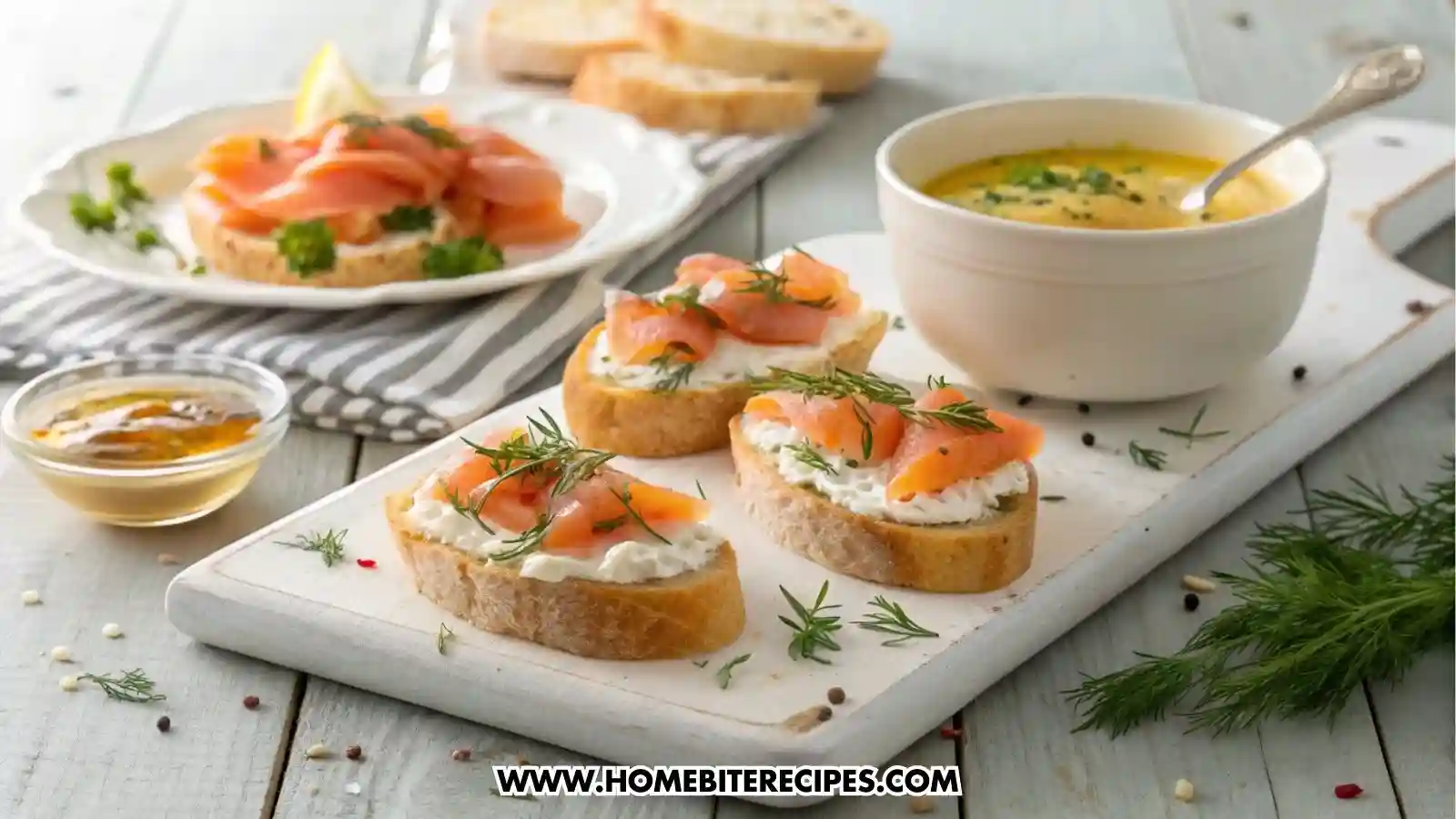 Easy smoked salmon crostini on a white serving board. Each crostini is topped with herbed cream cheese, folded slices of smoked salmon, fresh dill, and chives. Next to it, a bowl of warm, creamy potato soup garnished with parsley and a drizzle of olive oil.