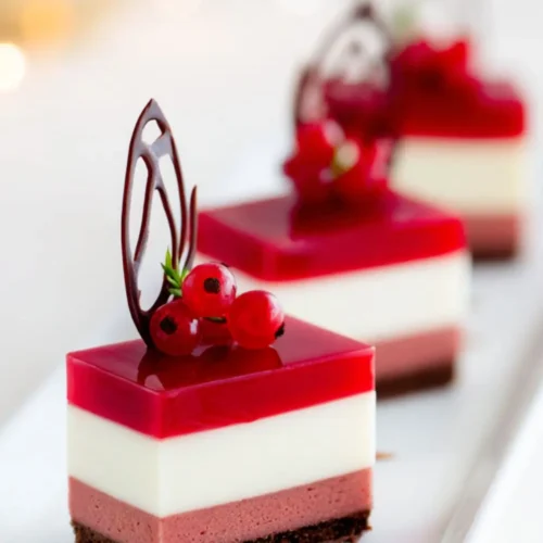 Elegant layered mousse cakes with chocolate base, strawberry mousse, vanilla cream, and glossy red berry jelly topping, decorated with chocolate art and currants
