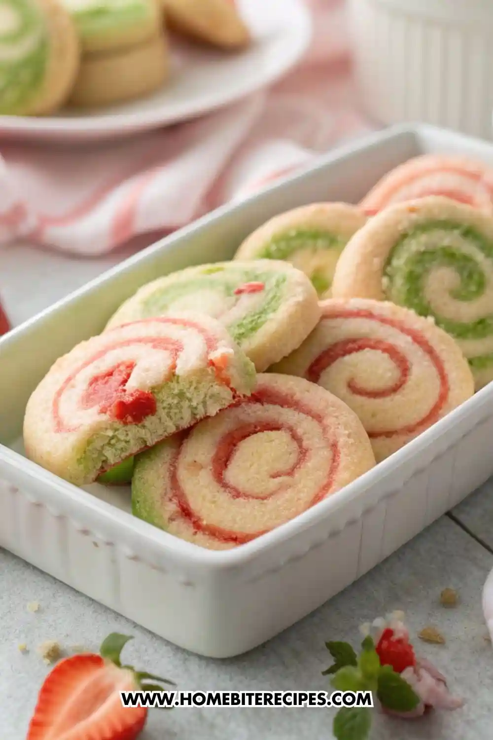 Colorful red and green pinwheel sugar cookies arranged in a white baking dish, with one cookie showing a bite revealing its soft texture.