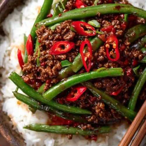 A close-up image of a spicy ground beef stir-fry with green beans and sliced red chili peppers served over fluffy white rice in a bowl with chopsticks.