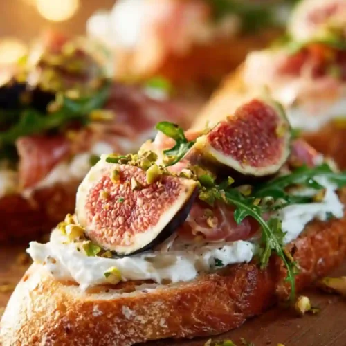 A close-up of gourmet crostini topped with whipped ricotta, prosciutto, fresh figs, arugula, and crushed pistachios on a wooden board.