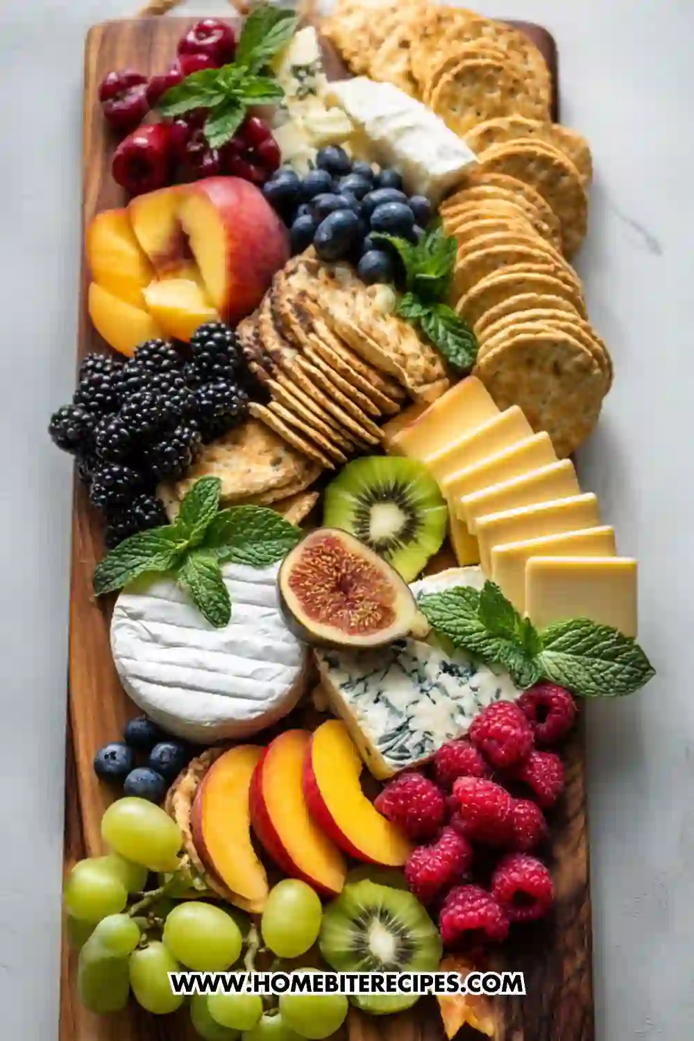 A colorful cheese and fruit charcuterie board featuring assorted cheeses, crackers, peaches, berries, figs, kiwi, and grapes beautifully arranged on a wooden serving platter with fresh mint garnish.