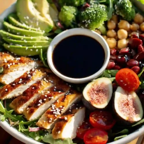 A colorful grilled chicken power bowl with avocado, figs, chickpeas, tomatoes, broccoli, and soy dressing served on a wooden table.