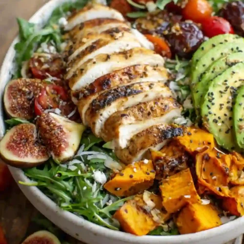 Colorful bowl of grilled chicken salad with avocado, roasted sweet potatoes, fresh figs, arugula, and cherry tomatoes topped with sesame seeds and balsamic drizzle.