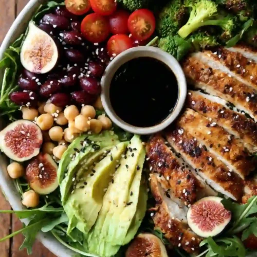 A colorful healthy chicken power bowl filled with sliced grilled chicken, avocado, figs, cherry tomatoes, chickpeas, kidney beans, broccoli, arugula, and a small bowl of balsamic dressing in the center.